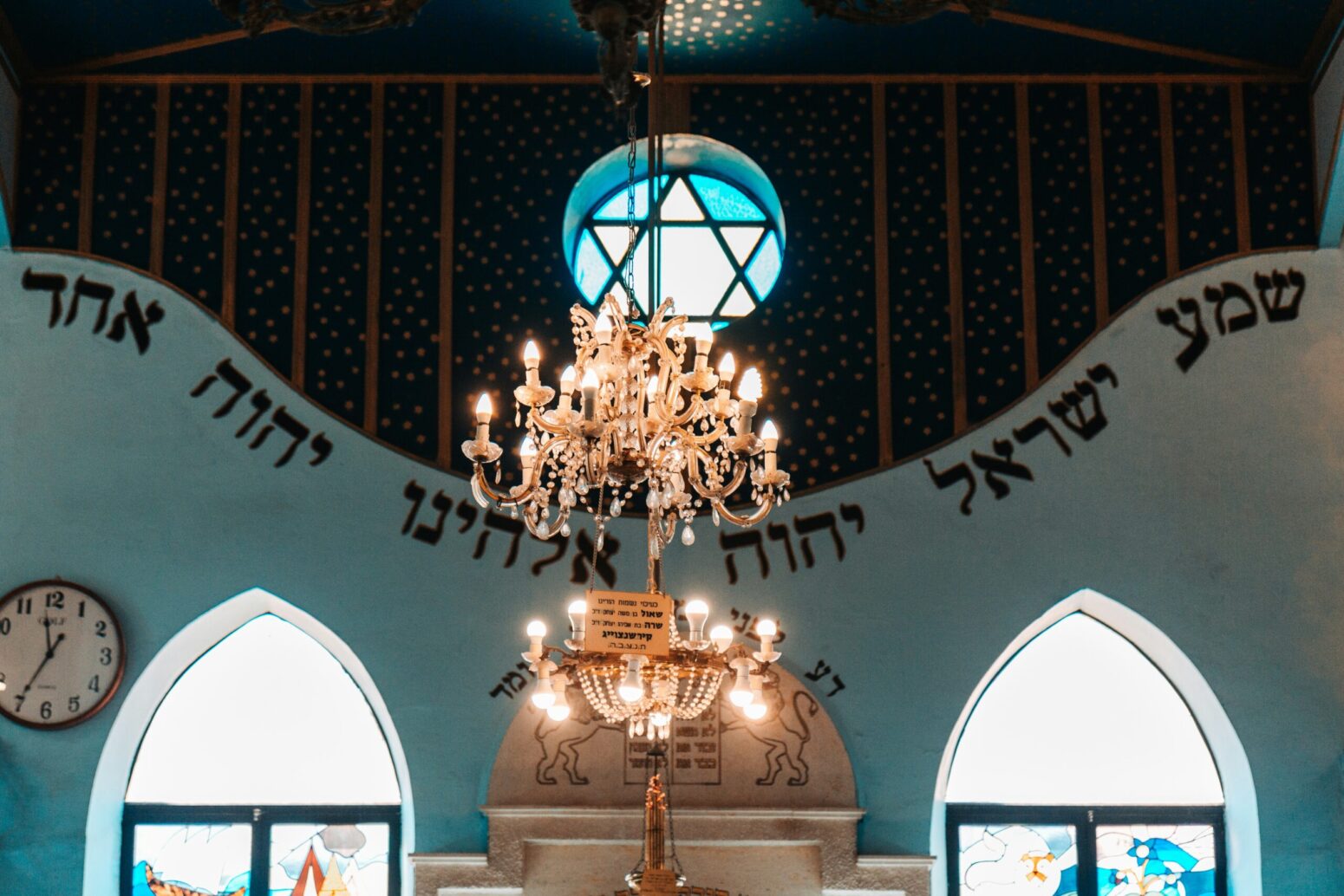 A chandelier hanging from the roof of a synagogue. There are three windows on the back wall, the middle of which, is a small, circular window with the star of David on it. The wall also has a prayer written in Hebrew.