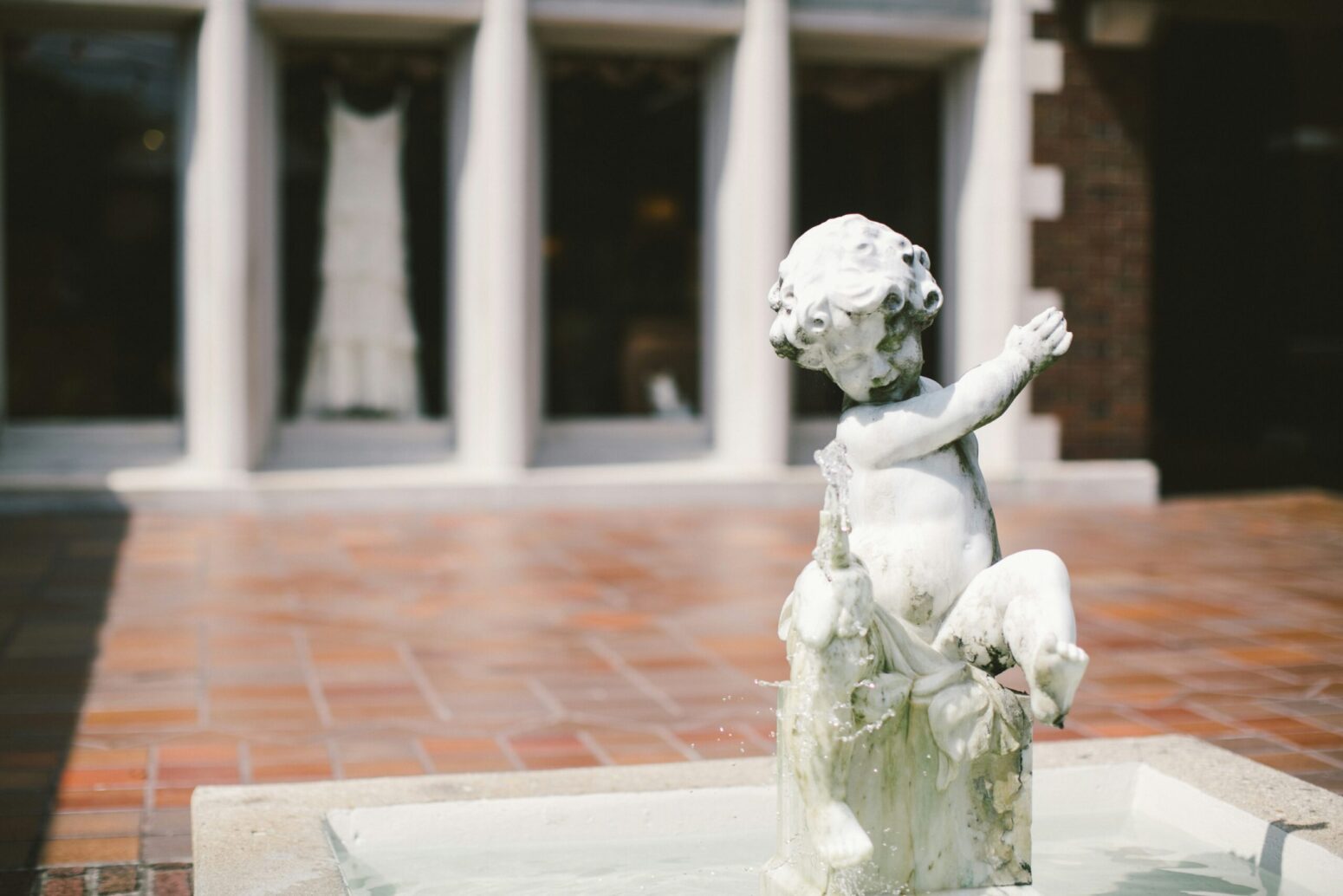 A small fountain in a brick courtyard. The center of the fountain has a small, marble statue of a child playing with a fish that spews water. In the background is a brick building with marble columns and large, wooden doors. A long, white, wedding dress hangs from one of the doors.