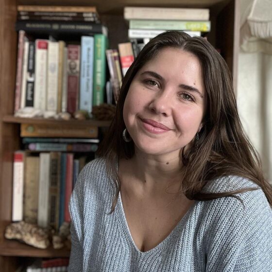 Mariah Rigg smiles in front of a bookshelf. She has long brown hair and wears earrings.