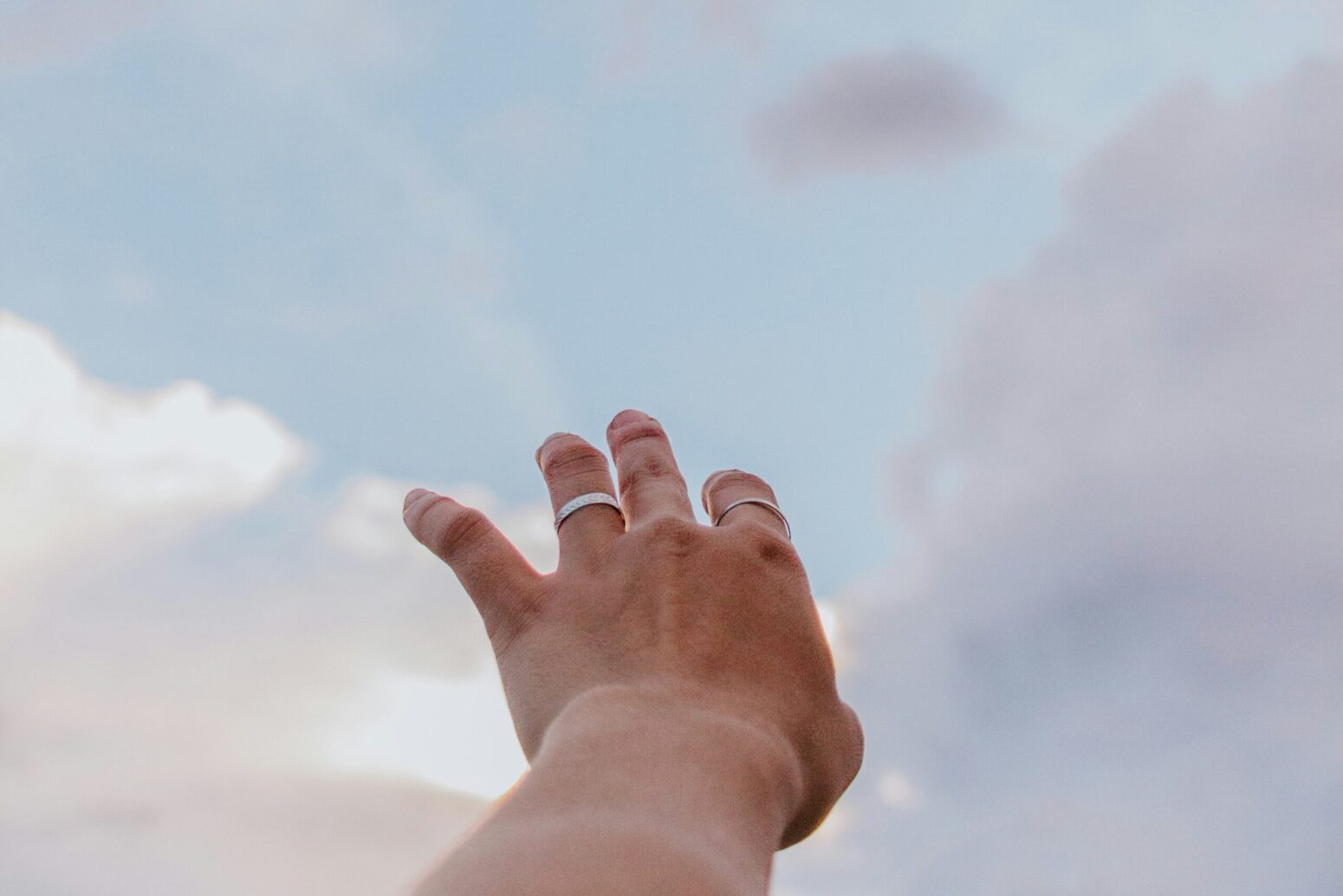 A perspective shot of someone reaching up with their left hand toward a blue, cloudy sky. The hand has two silver rings on it, one on the ring finger, and one on the pointer finger.