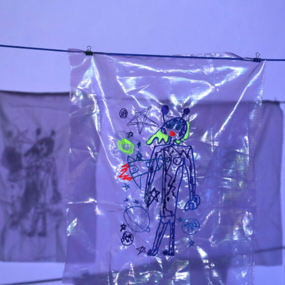 An embroidery of a child's drawing of an alien, spaceship, and planets on sheer, blue fabric, hung up on a clothesline.