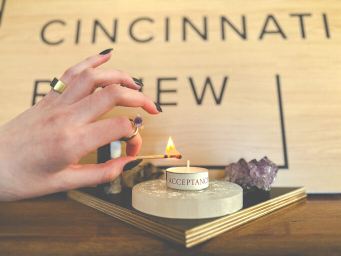A white hand with long black nails, wearing numerous rings, holds a lit match next to a tea light that reads, "Acceptance." It is placed on an altar of selenite, surrounded by crystals, in front of a woodgrain sign with the CR logo.