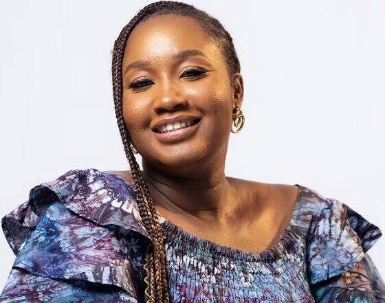A woman with light braids smiling. She sits in front of a white background wearing a purple-and-blue, ruffled dress.