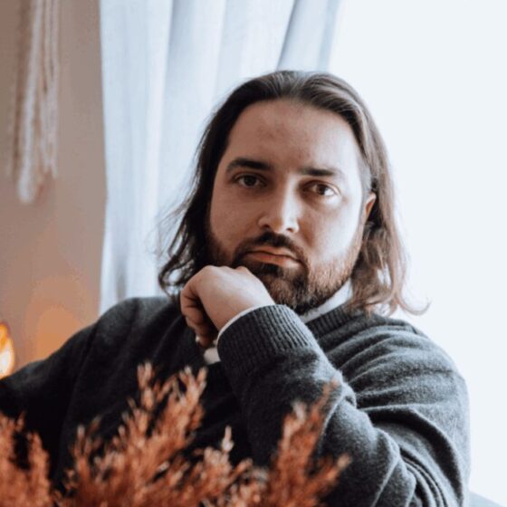 A man with straight, brown hair that goes down to his shoulders, a beard, and brown eyes. He sits in front of a window on a gray chair in a white room, wearing a dark gray sweater, resting his left elbow on the chair, and putting his hand to his chin. An orange, decorative plant slightly obscures the view.