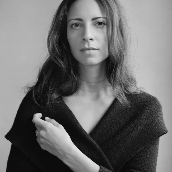 A grayscale photo of a woman with dark, straight hair that goes past her shoulders and dark eyes. She stands in front of a gray background, holding her left hand near her right shoulder. She wears a dark, V-neck dress with long sleeves.