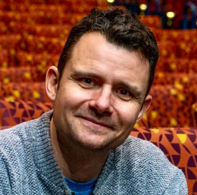 A man with short, brown, curly hair and brown eyes, smiling. He sits in an empty theater with red chairs, wearing a gray sweater