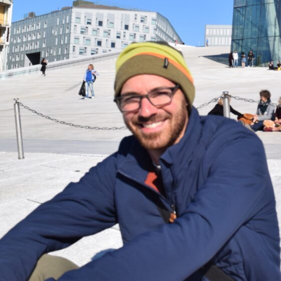 A man with a short beard, sitting, smiling, and looking at the camera. He wears glasses, a brown, yellow, and orange beanie, and a blue coat.