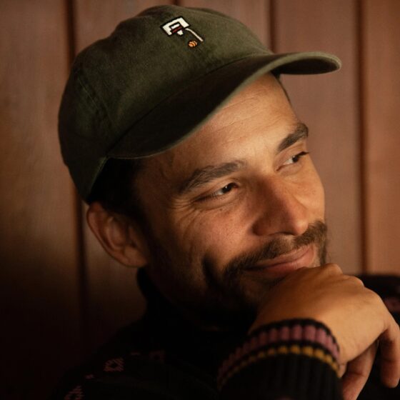 A man with a brown goatee and dark eyes, smiling. He sits in front of a wooden-panel background, wearing a green hat