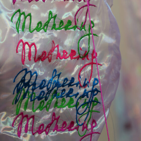 The word "Mothering" in a cursive font repeatedly embroidered onto a shiny, white piece of fabric. The words are in purple, pink, green, and blue, and they occasionally overlap.