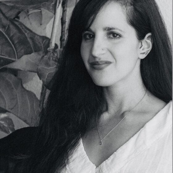 A grayscale photo of a woman with long, straight, dark hair and dark eyes, smiling and slightly facing left. She stands in front of a blank wall and a plant, and wears a lightly-colored shirt with a necklace.