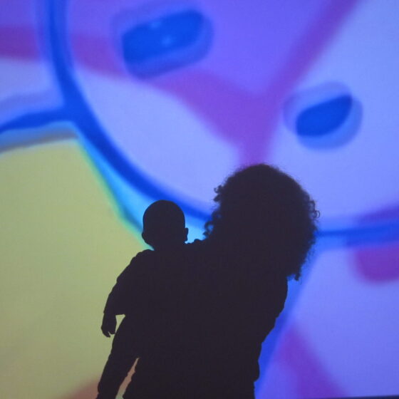 A silhouette of a woman with curly hair holding a baby boy, looking at a blue-and-yellow art projection.