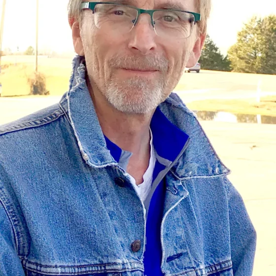 A man with short, brown hair and a gray beard, smiling. He wears rectangular glasses and a blue quarter-zip underneath a jean jacket.