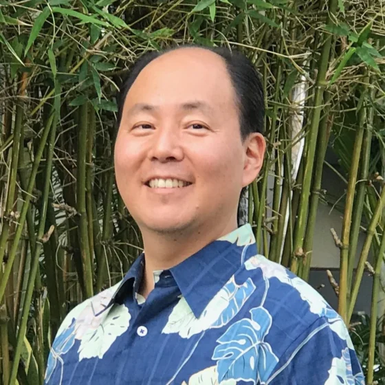 A man with short, black hair and brown eyes, smiling. He stands in front of bamboo, and wears a blue Hawaiian shirt