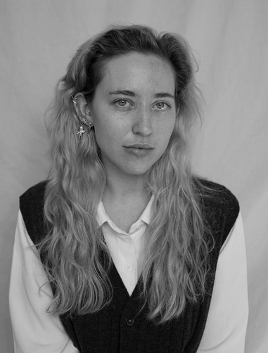 A grayscale photo of a woman with long, curly hair. She is looking at the camera, and wearing a collared, white shirt with a dark vest.