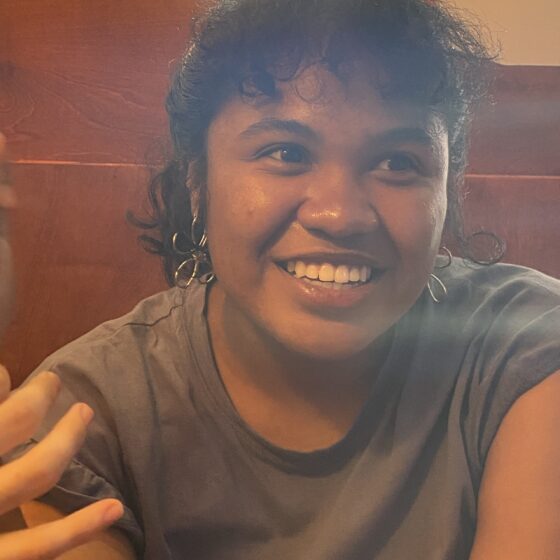 A Black and Asian American woman looking off to the side and smiling. She is wearing flower-shaped metal earrings and a gray T-shirt.