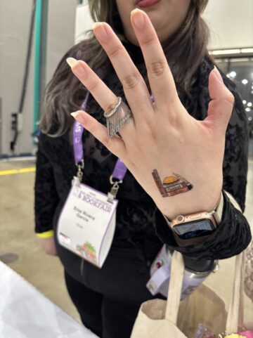 A woman with long nails wearing an ornate silver ring and Apple watch holds up the back of her hand to show where she's applied a temporary Cincinnati Review tattoo.