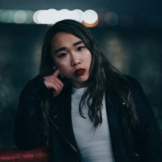 A woman with long, dark, wavy hair and dark eyes. She wears a white shirt, black leather jacket, and red lipstick.