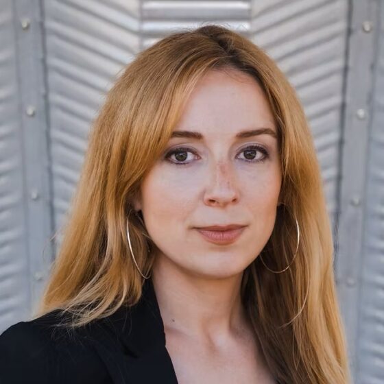 A woman with long, straight, red hair and brown eyes. She wears a black blazer and stands in front of a white, textured background