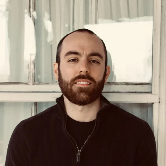 A man with a short, brown beard and a buzzcut, smiling. He leans against a window, wearing a black quarter-zip.