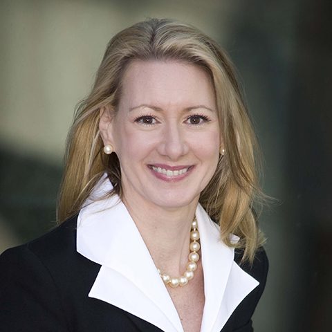 A woman with straight, blonde hair that goes past her shoulders and brown eyes, smiling. She wears a black blazer with a big, white collar on it, and a thick string of pearls around her neck.
