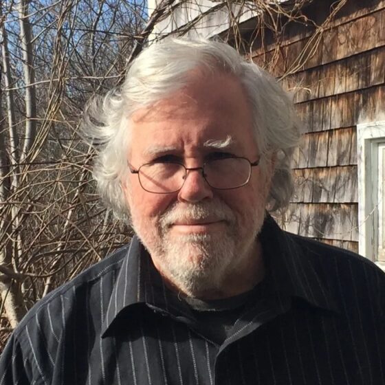 A man with white hair and a beard, wearing glasses and a black button-down shirt. He stands in front of an old building and a forest.