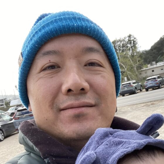 A man with a blue beanie, smiling, and holding a child wearing a blue hood with ears