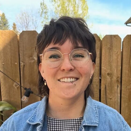 A woman with straight, brown hair in a mullet, blue eyes, and a lip ring, smiling. She stands in front of a wooden fence, and wears a black-and-white checkered shirt under a jean jacket.