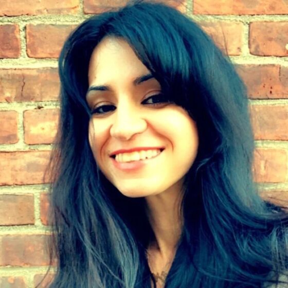 A woman with long, straight, black hair and dark eyes, smiling. She stands in front of a brick wall and wears a light green shirt