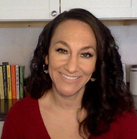 A woman with long, black, curly hair and brown eyes, smiling. She stands in front of a white cabinet with books on it, wearing a red shirt.