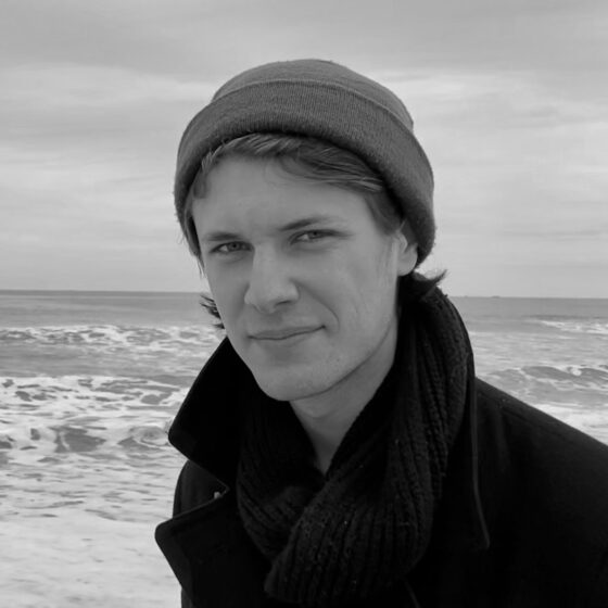 A black-and-white photo of Kylan Rice by a body of water with waves. Kylan looks directly at the camera, and is wearing a pea coat and beanie.