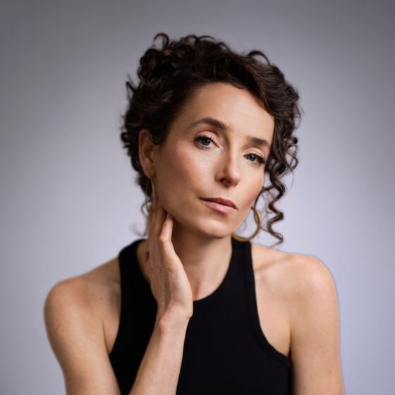 A woman with long, curly, brown hair tied up in a bun. She sits in front of a gray background, wearing a black tank top.