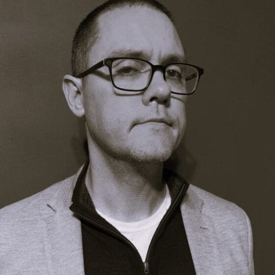 A grayscale photo of a man with dark, buzzed hair. He wears a white undershirt, a dark quarter-zip, a light-colored blazer, and square-shaped glasses