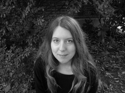 A grayscale photo of a woman with long, wavy hair, looking up at the camera. She sits in front of a bush, and wears a dark, long-sleeved shirt.