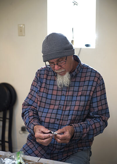 A man with a long, white goatee, looking down at a tube of paint. He sits in a room with white walls, in front of a wooden table with many different tubes of paint on it. He wears a gray beanie, black, rectangular glasses, and a red, blue, and black plaid shirt.
