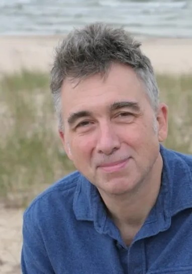 A man with short, gray hair, smiling. He sits outside in front of a beach, and wears a blue gold shirt.