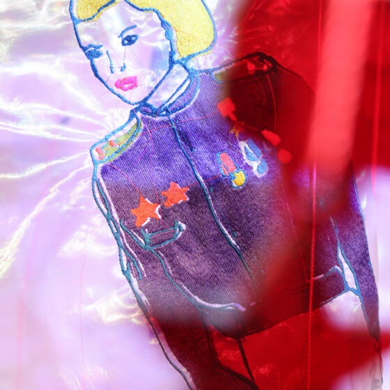 An embroidery of a blonde woman wearing a pilot's jacket, outlined in blue, on a white, iridescent piece of fabric.