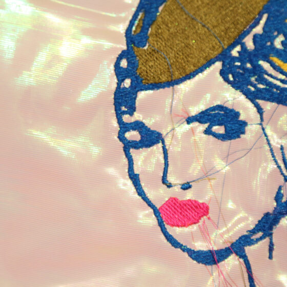 An embroidery of a woman with her hair in a bun, wearing a green pilot's hat, and outlined in blue, on a piece of white, iridescent fabric.