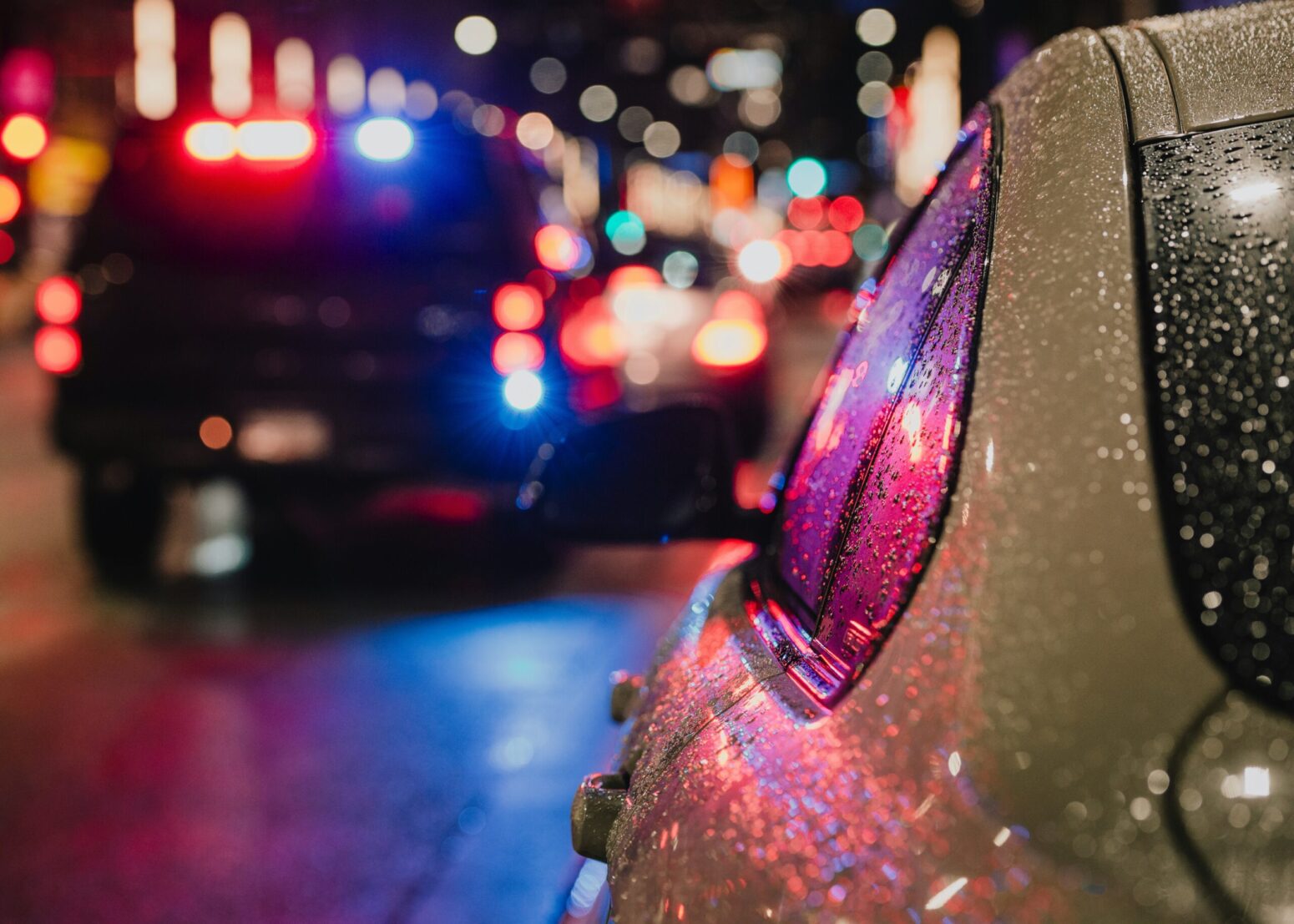 A close up view of the side of a car with a blurred city in the background. A police vehicle's lights glow, reflecting against the window