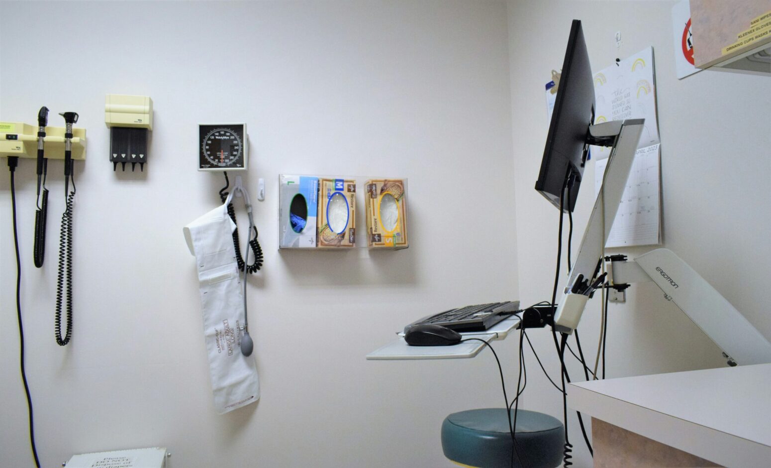 The inside of a doctor's office. Diagnostic tools, a scale, and boxes of gloves hang on the wall next to a computer.