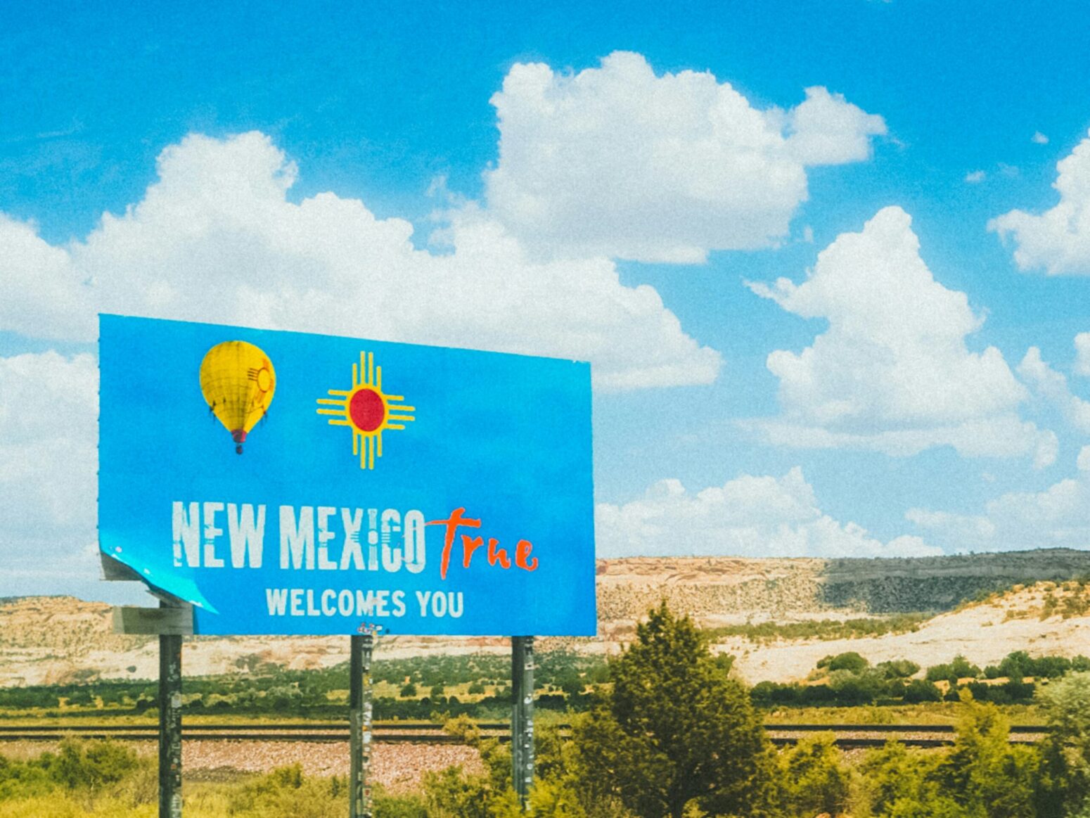 A blue road sign that reads "New Mexico True welcomes you" in white font with a picture of a yellow hot air balloon and the Zia Sun symbol overtop. The landscape behind the sign is flat, partially desert and partially plains, with a railroad track heading left to right.