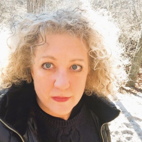 Selfie of Kelle Groom in the woods. She is wearing a jacket with the collar sitting under her curly hair, and there's a carpet of snow on the ground.