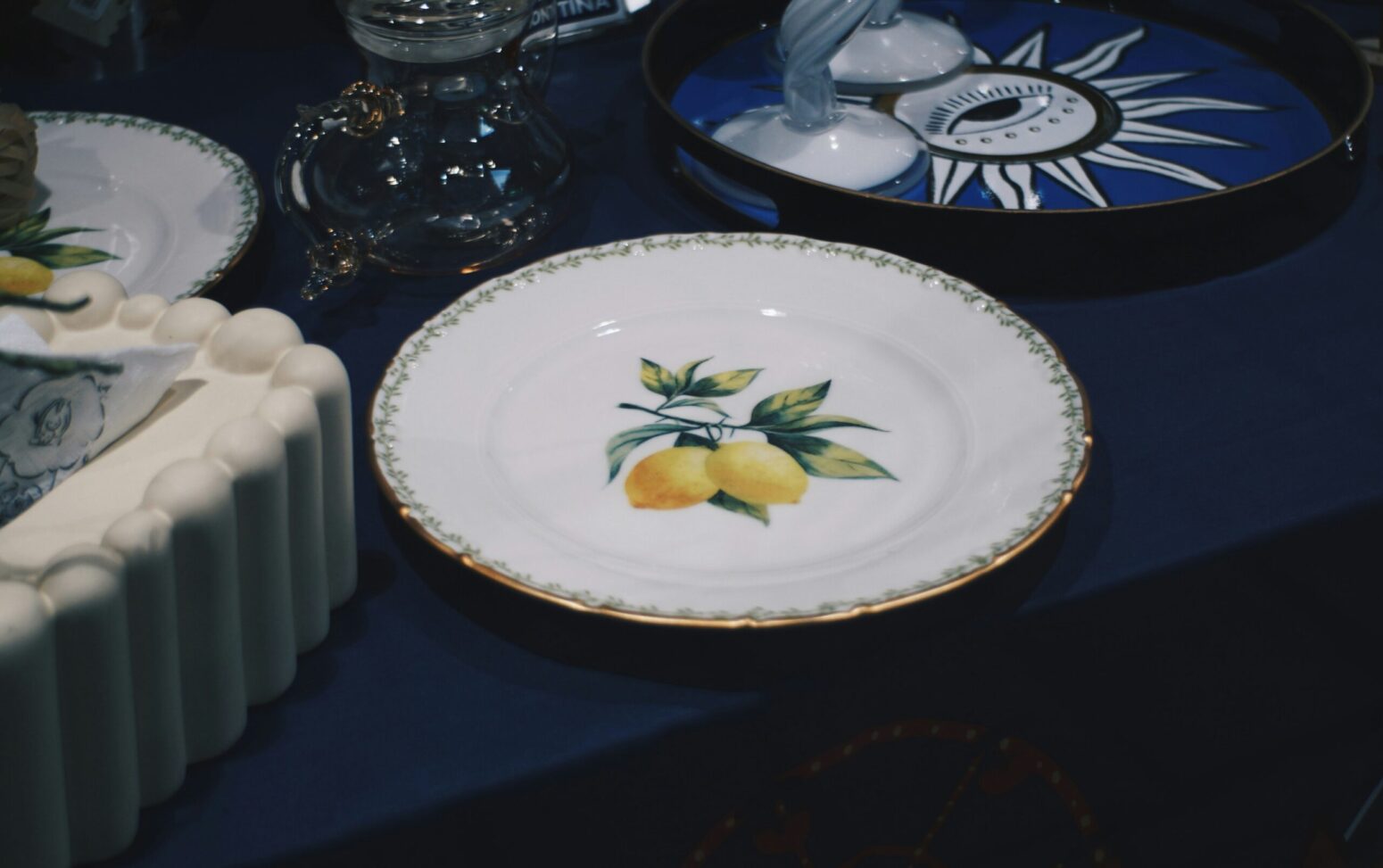 A fancy set of plates on a table with a blue tablecloth. The plate in the center of the frame is white with a golden border and a painted image of two lemons on a branch. To the left of the plate is a corner of a white-frosted cake. Above the cake is a white plate with a painted image of leaves in the center and a border that looks like vines. To the right of that plate is a round, glass pitcher. To the right of the pitcher is a blue tray with a white pattern of an eye in the middle of a sun on it. On the tray sits two white, glass goblets.
