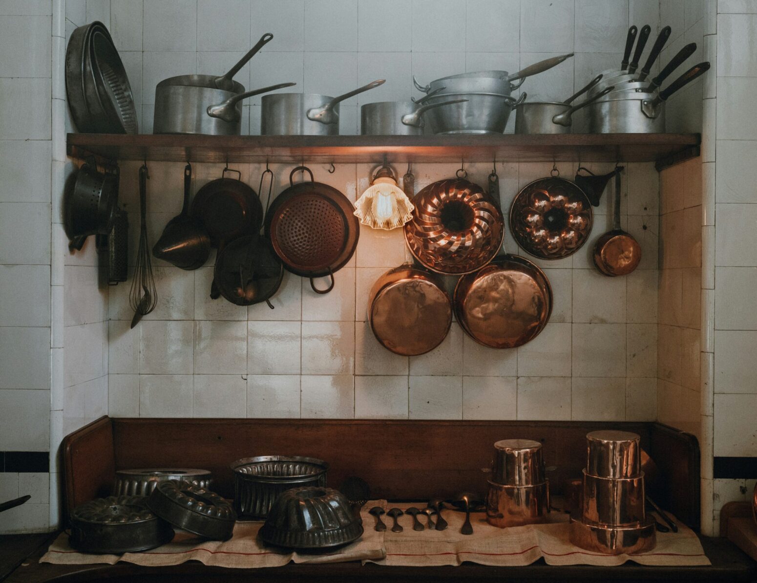 Silver and copper pots and pans hanging from or sitting on a shelf overtop a small table in a kitchen