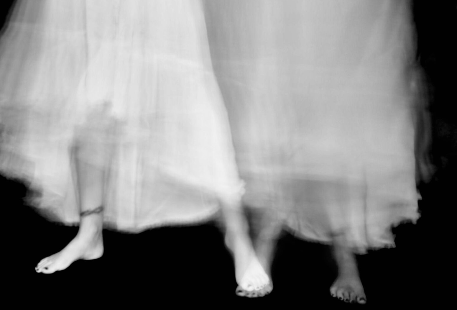 A black-and-white photo of the feet of two, blurry ghosts wearing long, white dresses.