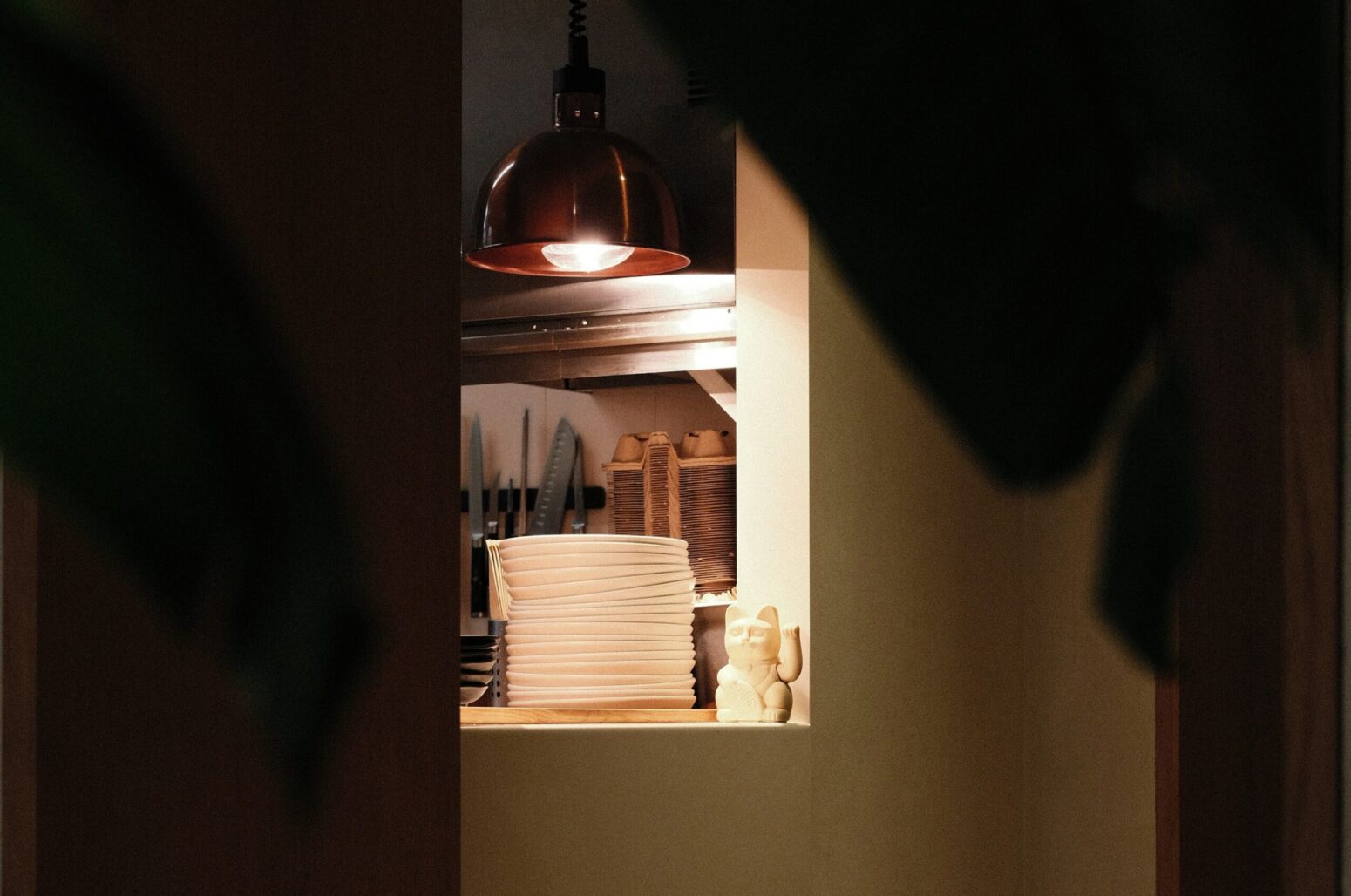An obscured view of a doorway to a kitchen, lit by a single light above the counter. A small, white, fortune cat sits on the right next to a stack of white plates. Behind them is a knife holder and a stack of cardboard drink holders.