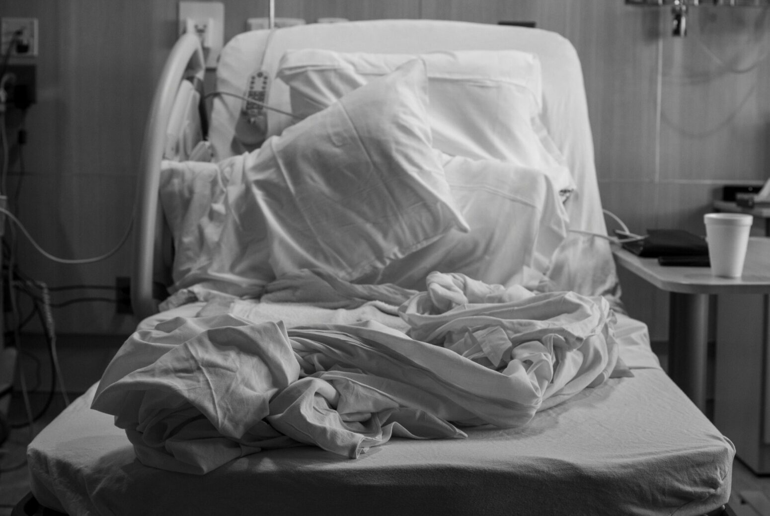 A grayscale photo of an empty hospital bed with disturbed pillows and sheets.