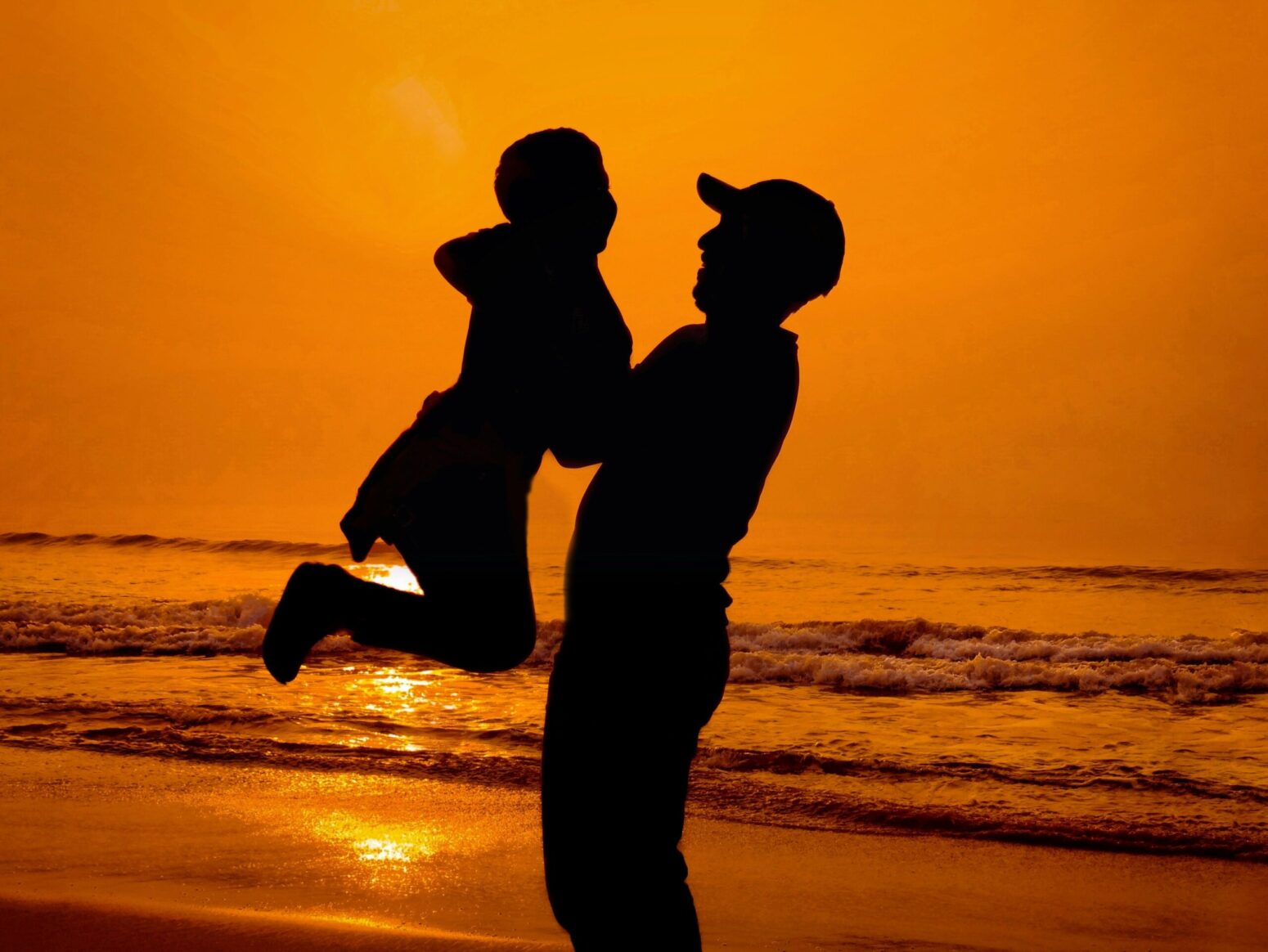 The silhouette of a man in a baseball cap holding up his son against an orange sunset on the ocean
