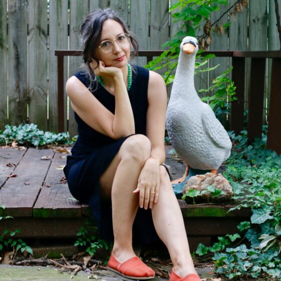 Danielle Dutton, a white woman in a black dress and red shoes, sits on a garden deck next to a goose statue.