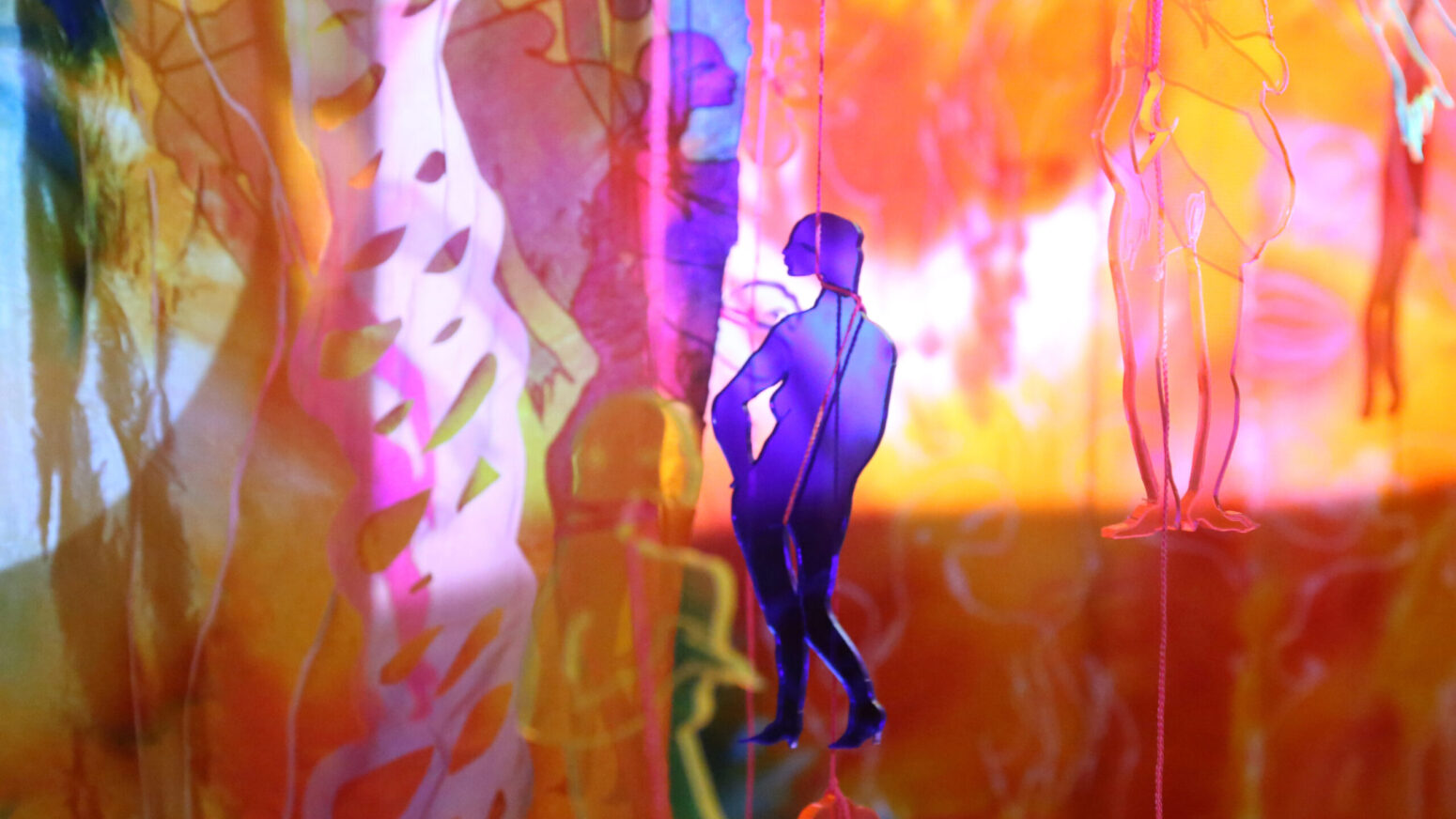 Multicolored, acrylic cutouts of naked pregnant women suspended from the ceiling by string. They hand in front of an orange-and-pink projection.