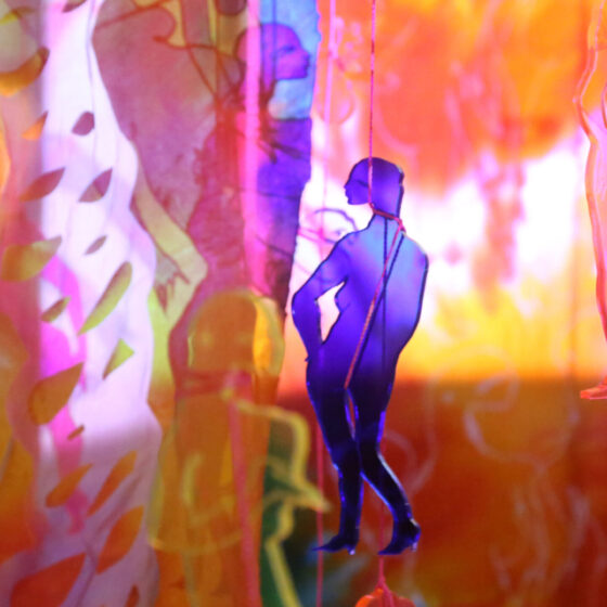 Multicolored, acrylic cutouts of naked pregnant women suspended from the ceiling by string. They hand in front of an orange-and-pink projection.